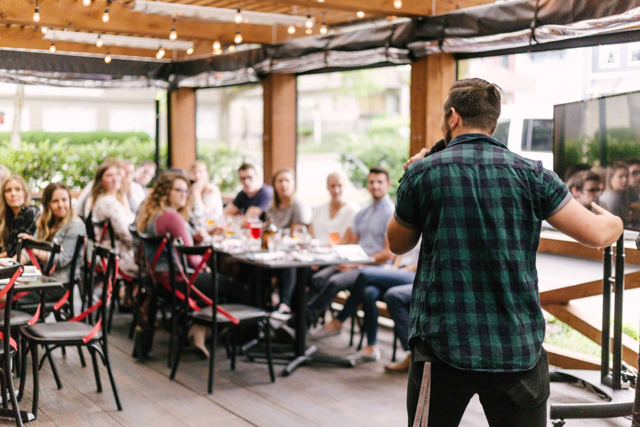 Someone speaking on a microphone at a meeting. The audience are sat at tables having eaten and drunk together and there are fairy lights above them on the wooden framed structure