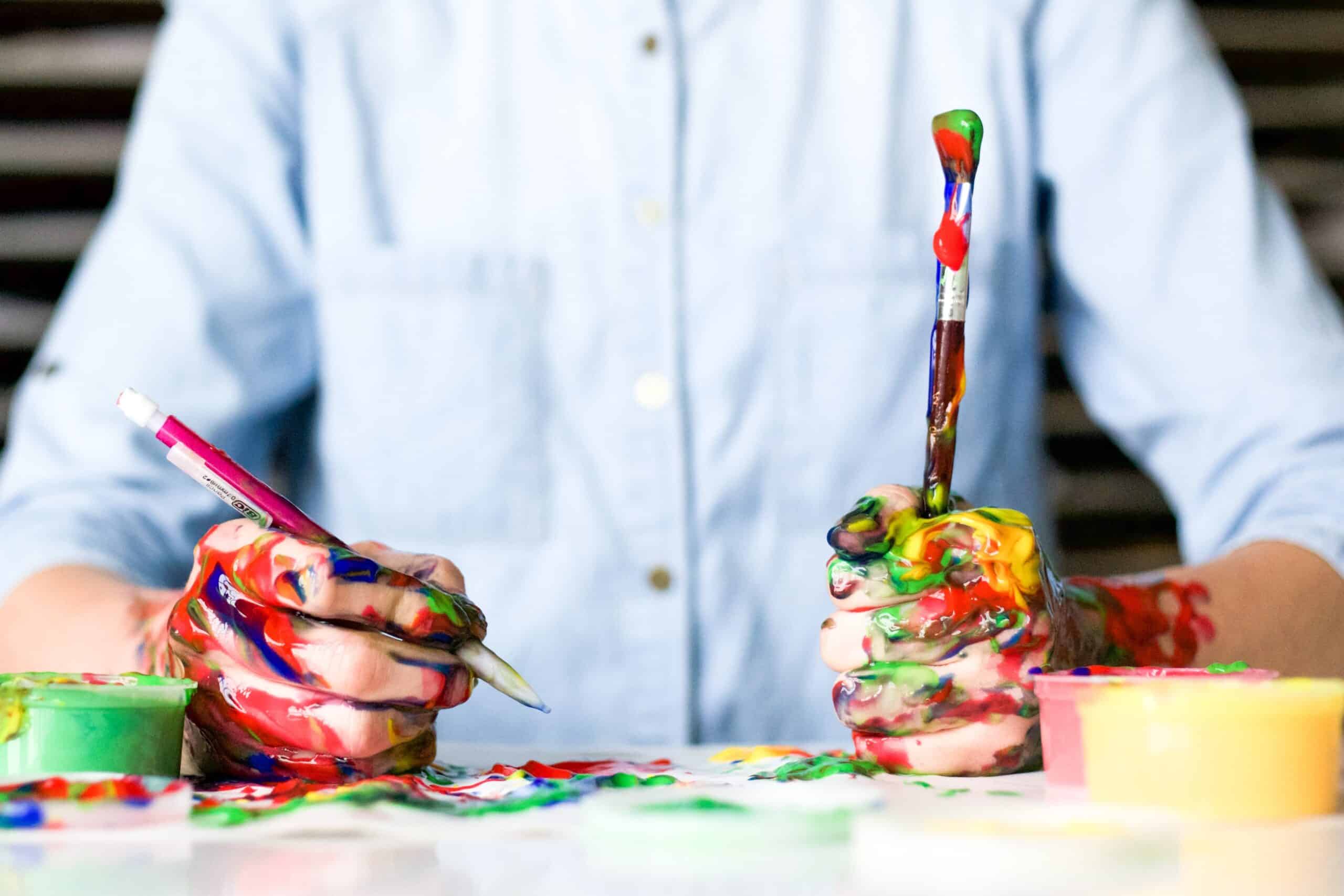 hands covered in runny, vibrantly-coloured paint, holding a brush and a pencil