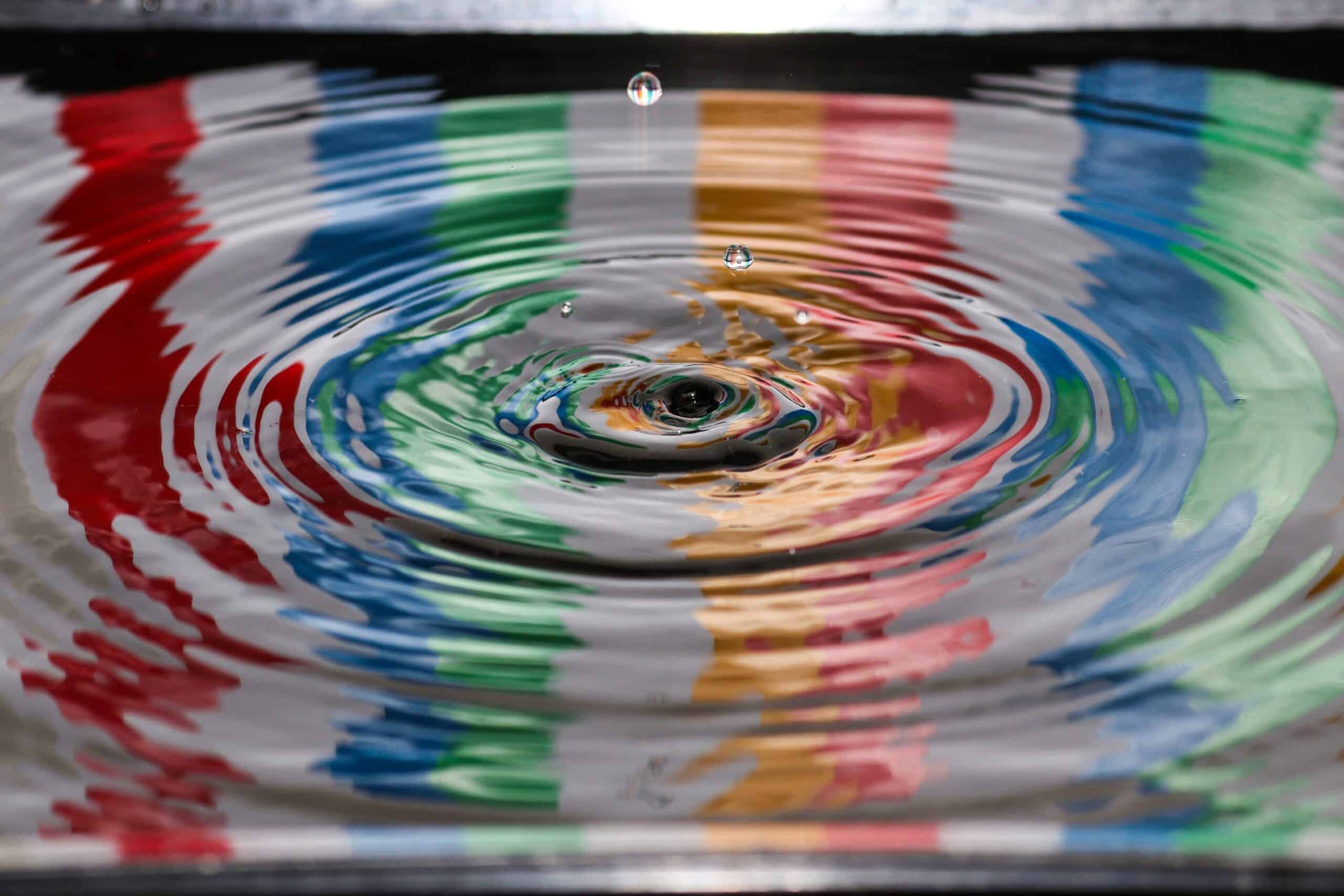 a drop of water bouncing off a large area of water with ripples, reflecting stripes of colour from an unseen source