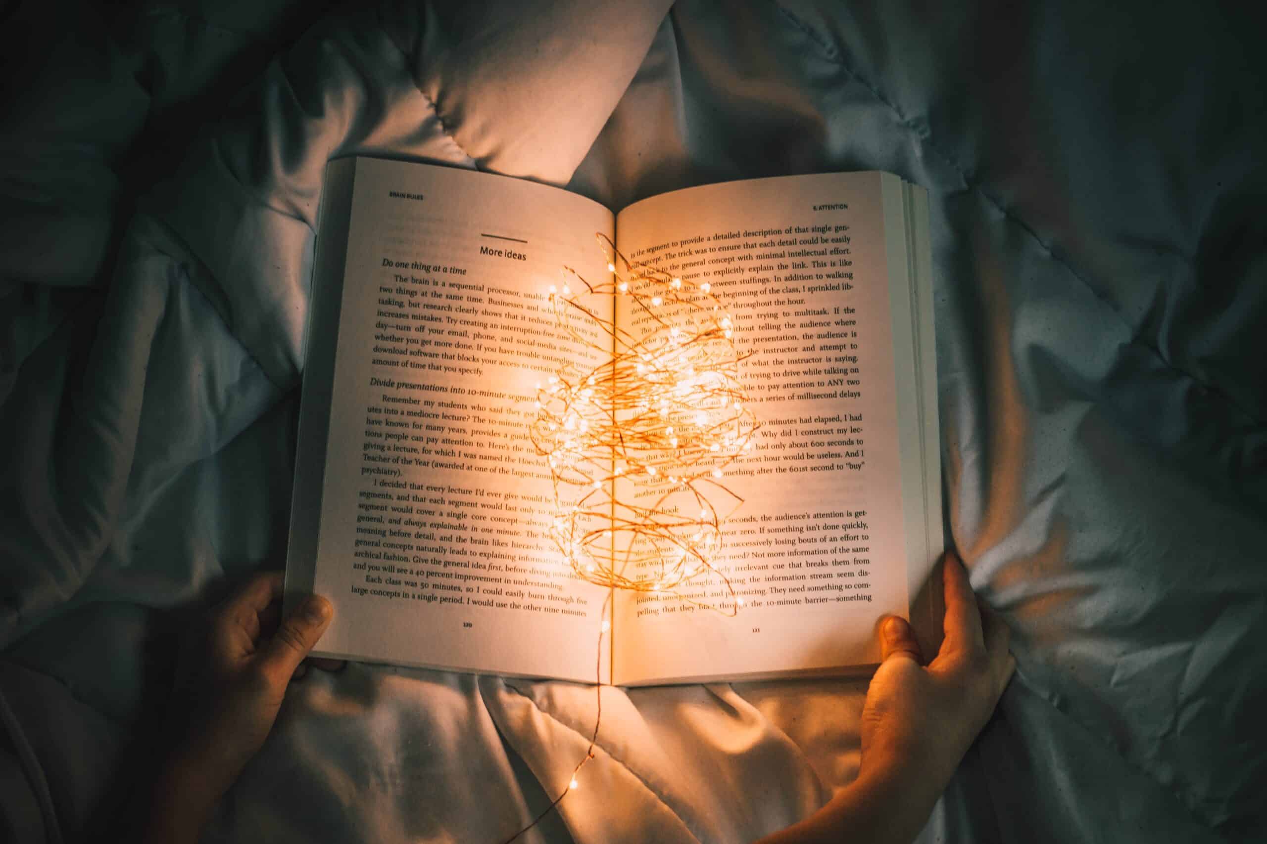 An open book on a bed at night time. The book is illuminated by small fairy lights.