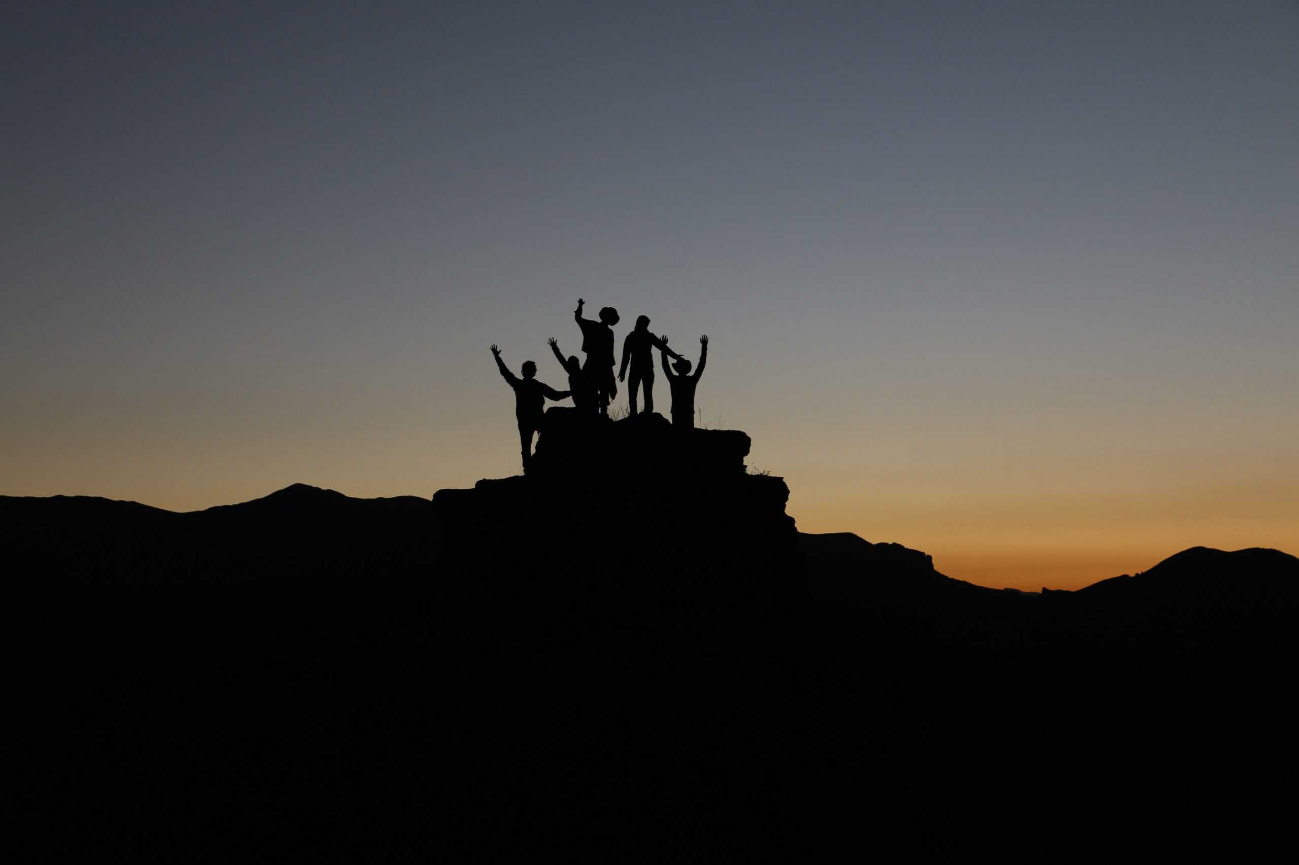the silhouette of a group of people celebrating on top of a rock, with a blue-gold sunset