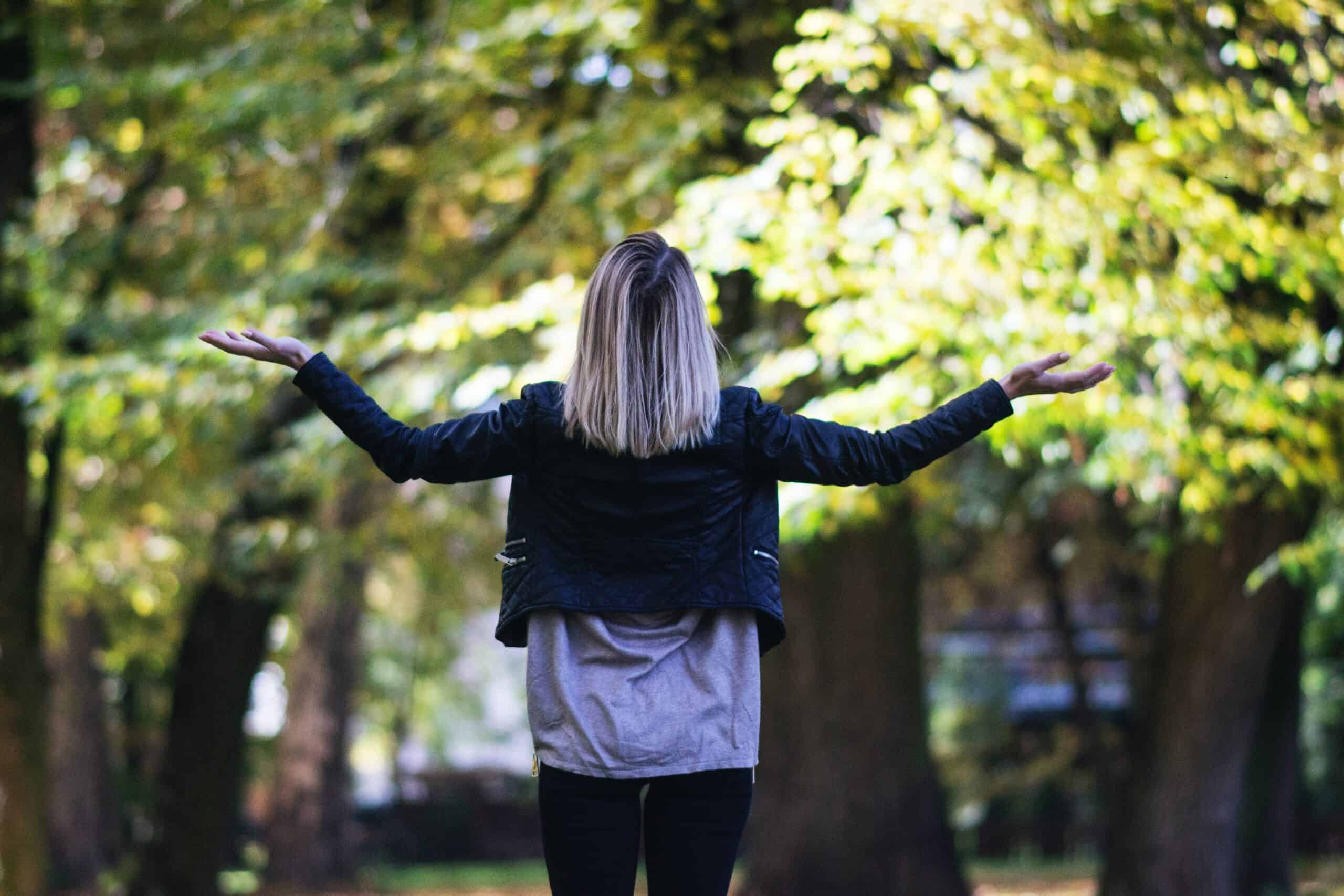 a woman with her hands stretched wide apart, palms towards the sun and trees that surround her, in a park