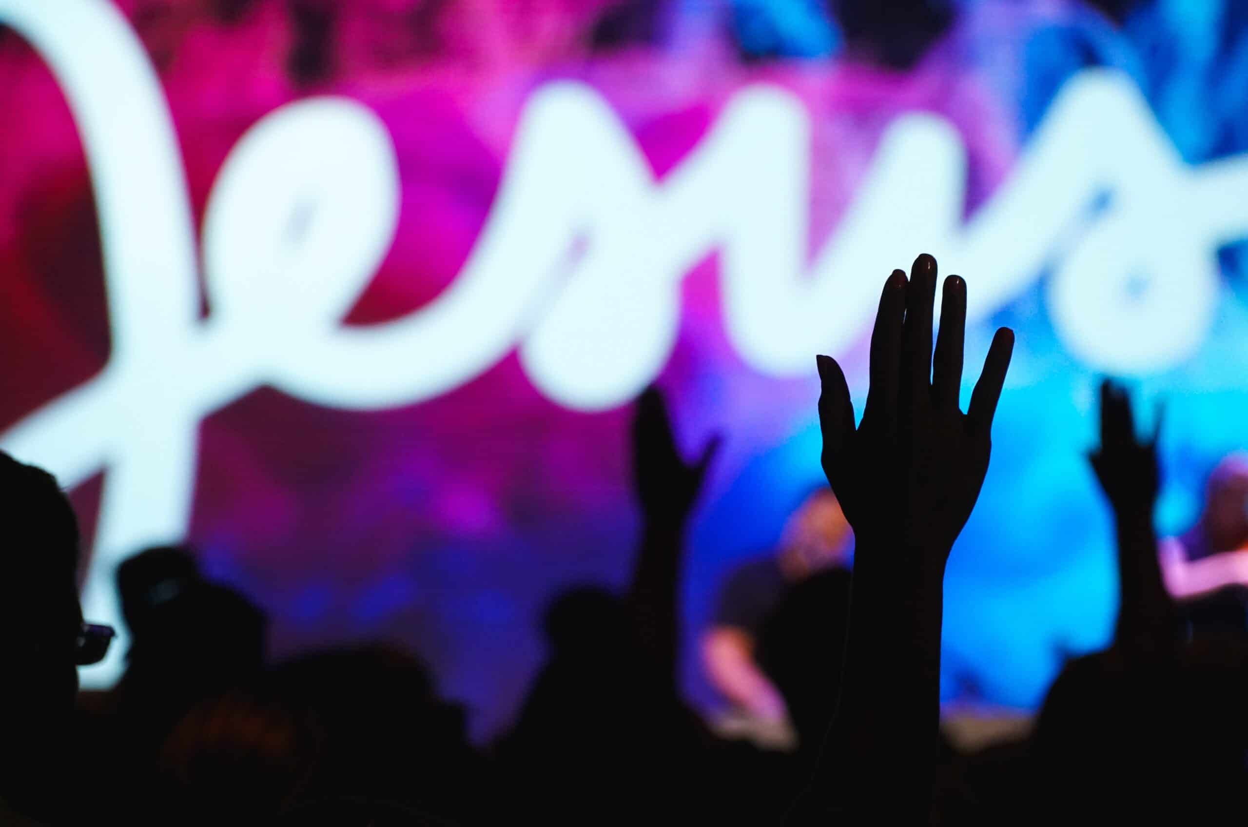 the silhouette of hands raised, with bright colours behind and the word 'Jesus' is illuminated